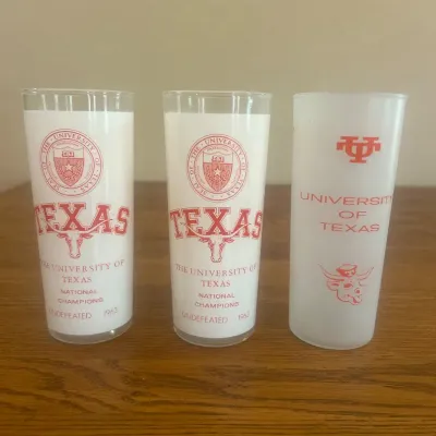 Sale Photo Thumbnail #269: This set includes three vintage University of Texas collector pint glasses featuring classic designs in orange. Two glasses display detailed portraits of notable figures associated with the university, while the third showcases the university logo along w