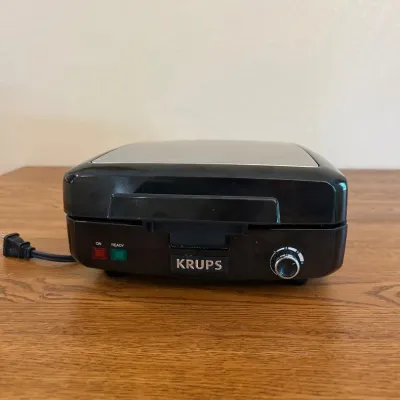 Sale Photo Thumbnail #240: This KRUPS waffle maker features a sleek black and stainless steel design, perfect for any kitchen countertop. It boasts four-section waffle plates for cooking multiple waffles at once. The compact appliance measures approximately 9.5 inches in width, 9 i