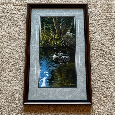 Sale Photo Thumbnail #82: Framed wildlife art print titled “Dancing Water – Loons” by Persis Clayton Weirs, depicting a pair of common loons floating on a quiet woodland pond. The vertical composition features finely detailed birds, reflective water, and a shaded forest back