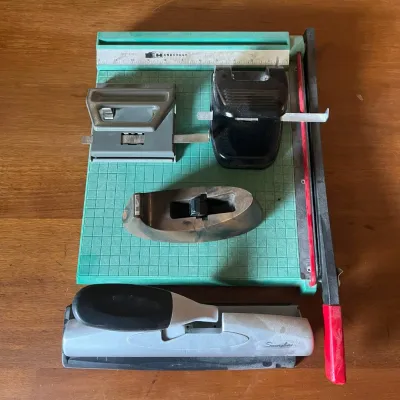 Sale Photo Thumbnail #303: This office set features a variety of essential desk tools including a sturdy paper cutter with a turquoise base and red safety handle, a black and gray dual hole puncher, a large gray hole puncher with a black rubber grip, and a unique wooden styled tape