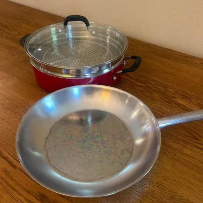 Sale Photo Thumbnail #231: This Tramontina cookware set includes a stainless steel frying pan and a red steamer pot with black handles and a glass lid. The frying pan features a polished stainless steel surface with a 12-inch diameter, offering excellent heat distribution and durab
