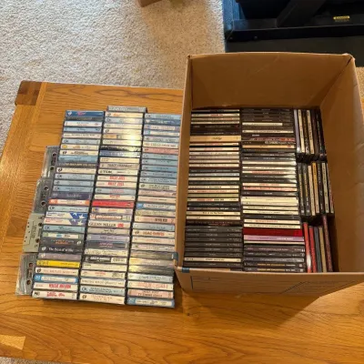 Sale Photo Thumbnail #216: This collection features a diverse assortment of compact discs and cassette tapes, neatly organized and stored in a cardboard box. The assortment includes various artists and genres, with cassettes showcasing vintage designs and labels. Highlighted aspect