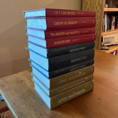 Sale Photo Thumbnail #312: This collection features eleven hardcover books showcasing works by William Faulkner, Ernest Hemingway, and John Steinbeck. The set is distinguished by its tri-color scheme: deep red for Faulkner's titles, navy blue for Hemingway's classics, and olive gre