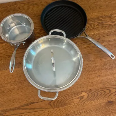 Sale Photo Thumbnail #230: This Calphalon 3-piece cookware set features a durable stainless steel saucepan, a grill pan with ribbed non-stick surface, and a large sauté pan with a tempered glass lid. Highlighted by its sleek, brushed metal handles and black non-stick interior, the