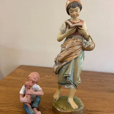 Sale Photo Thumbnail #285: This charming set of two child statues beautifully captures tender moments with exceptional detail. The first statue depicts a seated child holding a small dog, showcasing lifelike textures and earthy tones of muted white, blue, and brown. The second stat