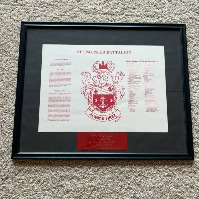 Sale Photo Thumbnail #55: This framed commemorative plaque honors the 1st Engineer Battalion, featuring the battalion's crest prominently in red and white with the motto 'Always First.' It includes detailed battalion history, battle honors, and three inscriptions: 'Grant of Arms,'