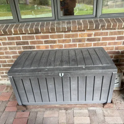 Sale Photo Thumbnail #213: This outdoor storage chest is designed for convenient garden tool and supply storage. It features a durable black plastic construction with a slatted panel design and is mounted on a dolly for easy mobility. Measuring 42 inches in width and 22.5 inches in