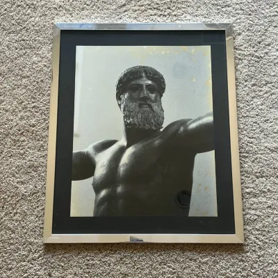 Sale Photo Thumbnail #86: This framed black and white photograph features a detailed close-up of a classical male statue showcasing muscular torso and bearded face with an outstretched arm. The piece is highlighted by a sleek silver metallic frame that adds a modern touch to the c