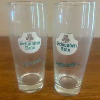 Sale Photo Thumbnail #268: This set features two Schwaben Bräu branded pint glasses, distinguished by their clear glass material and iconic logo printed on the front. Each glass stands approximately 6.75 inches tall with a diameter of about 3 inches at the top, making them ideal f