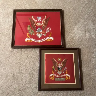 Sale Photo Thumbnail #50: This set features two beautifully framed embroidered crests representing the Twenty-Third Engineers and the First Engineers units. Each crest is meticulously stitched on a vibrant red fabric background, highlighting the detailed eagle insignia and unit ba