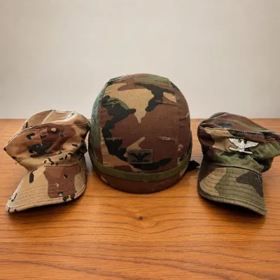 Sale Photo Thumbnail #60: Three-piece group of U.S. military camouflage headgear from the collection of a former U.S. Army battalion commander. Includes a woodland camouflage-covered combat helmet, a six-color desert “chocolate chip” camouflage patrol cap, and a woodland camou