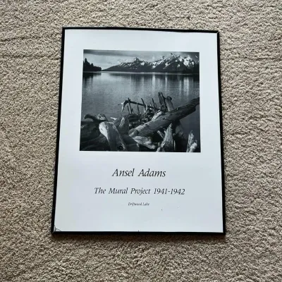 Sale Photo Thumbnail #116: This framed black and white photographic print features Ansel Adams' iconic image from The Mural Project 1941-1942, titled 'Driftwood Lake.' The piece highlights Adams' masterful landscape photography capturing serene natural scenery with driftwood in the