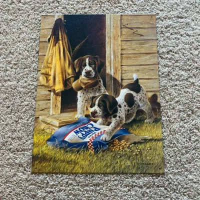 Sale Photo Thumbnail #100: This metal wall art, created by artist Lynn R. Kaatz in 1997, showcases two playful spotted puppies interacting with a spilled bag of 'Puppy Chow' dog food outside a rustic wooden shed. The piece highlights vivid earthy tones with detailed grass textures 
