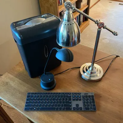 Sale Photo Thumbnail #316: This office set features a powerful black paper shredder with a separate waste bin, a slim modern black and silver wired keyboard, and two versatile desk lamps. The first lamp is a sleek black design with a curved neck and dome-shaped shade, ideal for foc