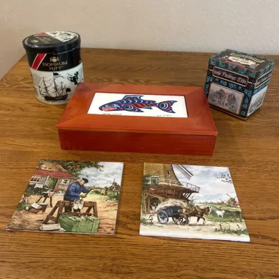 Sale Photo Thumbnail #288: This unique collection of various collectors' items includes a wooden box with a colorful fish design, two vintage-style decorative tins with intricate artwork, and two detailed trivets depicting rural scenes. The wooden box is crafted from warm-toned woo