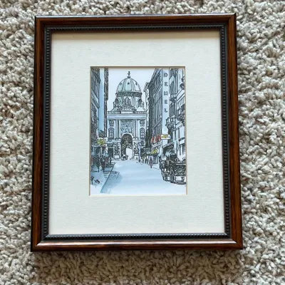 Sale Photo Thumbnail #119: This framed art captures a detailed architectural street scene, featuring a prominent domed building in the background. The artwork highlights fine line work and subtle color accents that bring the cityscape to life. The piece is set within a dark wood fr