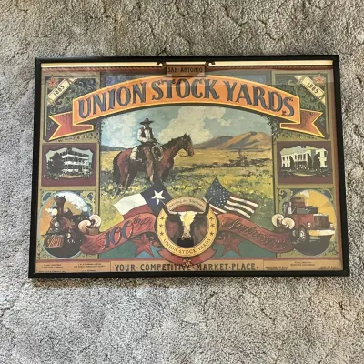 Sale Photo Thumbnail #108: Framed commemorative poster celebrating 100 years of the Union Stock Yards in San Antonio, Texas, 1889–1989. The colorful Western-themed artwork features a mounted cowboy, Texas and American flags, longhorn imagery, a steam locomotive, semi truck, and h