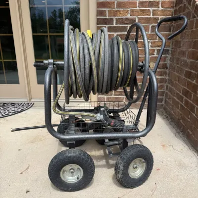 Sale Photo Thumbnail #211: This portable hose caddy features a robust metal frame with sturdy wheels for easy maneuverability. It includes a long black hose neatly coiled on the reel and a durable spray nozzle with multiple settings. The hose caddy is designed for convenient outdoo