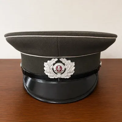 Sale Photo Thumbnail #13: Original Cold War-era East German National People’s Army (NVA) visor cap, featuring a field-gray/olive body with light piping, glossy black visor and black chinstrap. The front displays the distinctive DDR national emblem with hammer and compass surroun