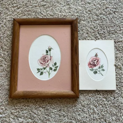 Sale Photo Thumbnail #123: This set features two delicate hand-painted watercolor illustrations of roses, each framed individually. One piece showcases a pink rose with rich green leaves set against a soft pink mat, enclosed in a warm-toned wooden frame. The second painting, displa