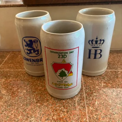 Sale Photo Thumbnail #298: This unique set of 3 stoneware beer steins includes distinct designs from Lowenbrau, HB, and a special Organization Day 23rd enamel insignia from 1964. Each stein stands 7.5 inches tall and measures 3.5 inches wide, crafted from durable stoneware with tra