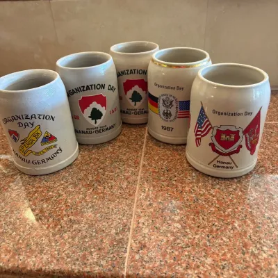 Sale Photo Thumbnail #301: This collection features 5 vintage beer steins commemorating 'Organization Day' events held by United States Army units stationed in Hanau, Germany. Each stein is crafted from sturdy ceramic material with a traditional stein shape, showcasing detailed mil