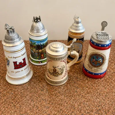 Sale Photo Thumbnail #297: This collection features a set of five authentic German-themed beer steins, each uniquely decorated with detailed artwork and traditional motifs. The steins are crafted from ceramic material with intricate painted designs showcasing rural and hunting scen