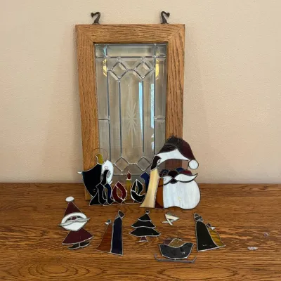 Sale Photo Thumbnail #329: This set features a collection of stained glass Christmas decorations depicting a nativity scene, Santa figures, a Christmas tree, a sleigh, and the word 'Noel'. Each piece is crafted from colorful stained glass with black soldered outlines, offering a fe