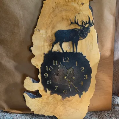 Rustic live-edge wood wall clock with a metal laser-engraved elk silhouette and a textured clock face with maple leaf detail
