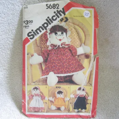 Sale Photo Thumbnail #24: Vintage Simplicity sewing pattern number 5682 for a stuffed rag doll with various outfit options. One size. Includes transfers for embroidery. The envelope shows wear, including creasing, edge tears, and a small sticker on the front. Pattern pieces and in