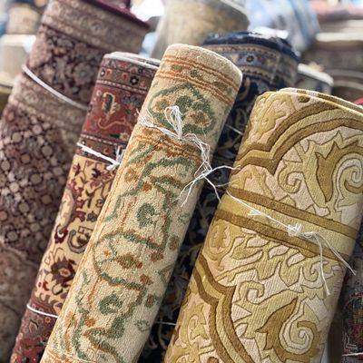Close-up assortment of rolled rugs showcasing blue, gold, ivory, green, and neutral color palettes with detailed floral and decorative...