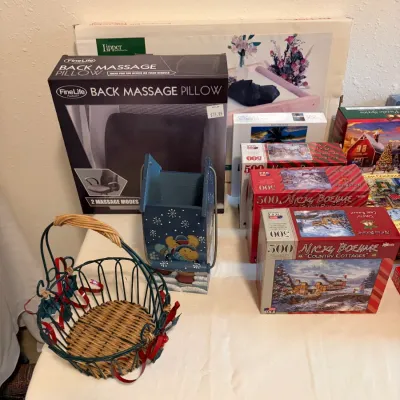 Sale Photo Thumbnail #80: Assorted home goods and activities, including a boxed FineLife back massage pillow, several Nicky Boehme jigsaw puzzles, a decorative winter-themed wooden box, and a wire basket with a woven wicker bottom.