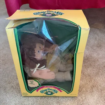 Sale Photo Thumbnail #96: Vintage 1980s Coleco Cabbage Patch Kids doll in its original box, featuring brown hair and glasses. The yellow box shows some wear and creasing on the top flap.