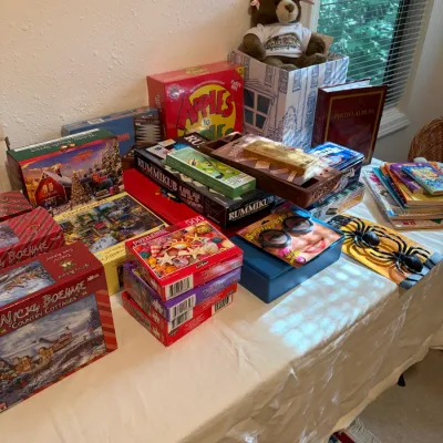 Sale Photo Thumbnail #81: Assortment of family board games, jigsaw puzzles, and novelty items, including Apples to Apples, Rummikub, and Nicky Boehme puzzles, arranged on a table alongside a plush teddy bear and books.