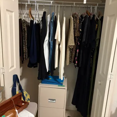 Sale Photo Thumbnail #31: Assorted women's clothing hanging in a closet, featuring dresses, blouses, and knitwear, alongside a two-drawer metal filing cabinet.