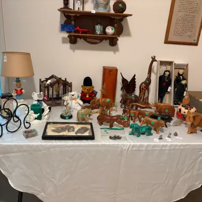 Sale Photo Thumbnail #28: A collection of vintage collectibles displayed on a table, featuring numerous hand-carved wooden animal figurines, boxed porcelain dolls, stuffed animals, and a table lamp.