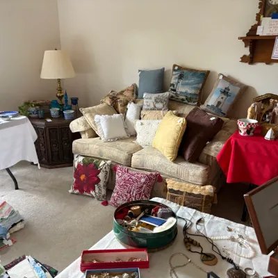 Sale Photo Thumbnail #12: An assortment of home goods, featuring a beige upholstered sofa piled with decorative throw pillows, tables displaying glassware and holiday decor, and a selection of jewelry and linens in the foreground.