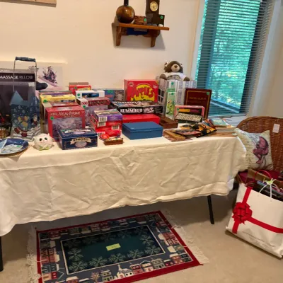 Sale Photo Thumbnail #29: Assortment of board games, puzzles, and home goods, including Apples to Apples, Rummikub, and a back massage pillow, displayed on a table alongside a wicker armchair.