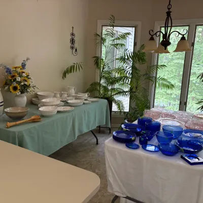 Sale Photo Thumbnail #20: Collection of vintage tableware featuring white ceramic dinnerware and a stoneware crock on one table, alongside cobalt blue glassware and pink depression glass on an adjacent table.