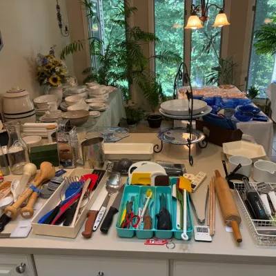 Sale Photo Thumbnail #87: A large assortment of kitchenware and household items, including utensils, baking tools, ceramic dishes, and glassware, displayed on a counter and tables for an estate sale.