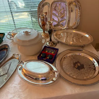 Sale Photo Thumbnail #40: Collection of vintage silver-plated serving ware, including tree-and-well platters, a covered casserole dish, and an engraved round tray, displayed alongside a white ceramic soup tureen and candlesticks.