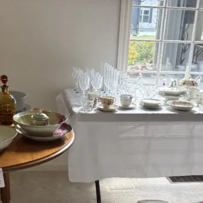 Sale Photo Thumbnail #11: Collection of vintage tableware and glassware, featuring stacked ceramic plates, mugs, an amber glass decanter, delicate teacups, saucers, and crystal stemware displayed on tables.