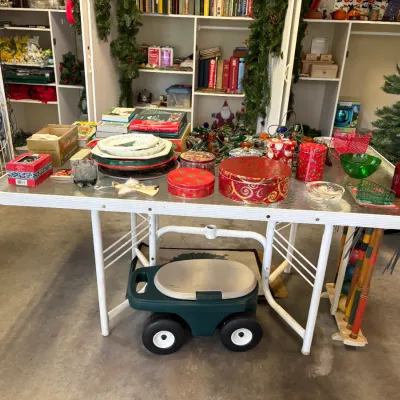 Sale Photo Thumbnail #4: White metal table with a textured glass top, displaying an assortment of Christmas tins, plates, and holiday decor, with a rolling garden seat stored underneath.