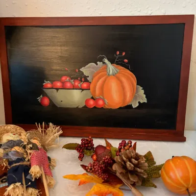 Sale Photo Thumbnail #92: Framed still life painting depicting a pumpkin and a bowl of apples on a dark background, displayed alongside autumn decorations including miniature scarecrows and faux foliage.