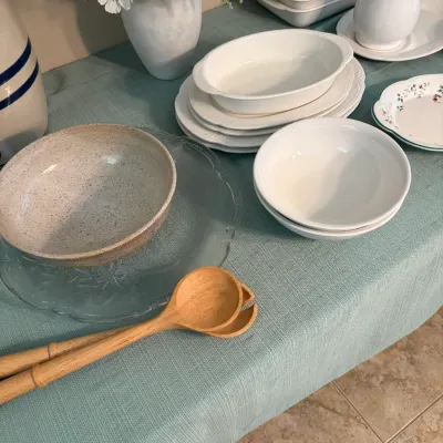 Sale Photo Thumbnail #63: Assorted farmhouse-style tableware including a speckled ceramic bowl on a clear glass platter, wooden serving utensils, and various white ceramic serving dishes and pitchers.