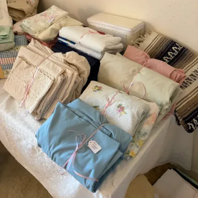 Sale Photo Thumbnail #36: Assorted vintage bed linens, sheets, and blankets tied with pink ribbons on a display table. The collection includes floral patterns, solid pastels, lace-trimmed pieces, and a woven serape blanket.