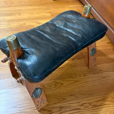 Sale Photo Thumbnail #70: Vintage camel saddle stool featuring a black tooled leather cushion on a wooden frame with brass-capped posts and decorative metal studs.