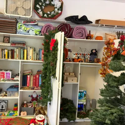Sale Photo Thumbnail #3: White storage shelving units filled with holiday decorations, books, rolled sleeping bags, and household goods, featuring artificial Christmas trees, garlands, and Halloween decor.