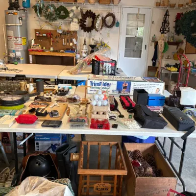 Sale Photo Thumbnail #6: Garage featuring folding tables filled with miscellaneous household items, including glassware, golf balls, a boxed TrimRider exercise machine, and vintage wooden crates.