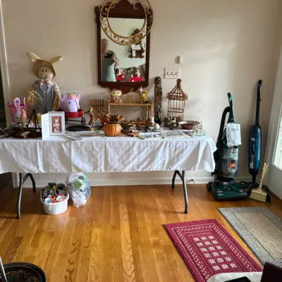Sale Photo Thumbnail #15: A folding table displaying an assortment of home decor, including Easter decorations, a wicker birdcage, and ceramic figurines, flanked by two upright vacuum cleaners on a hardwood floor.